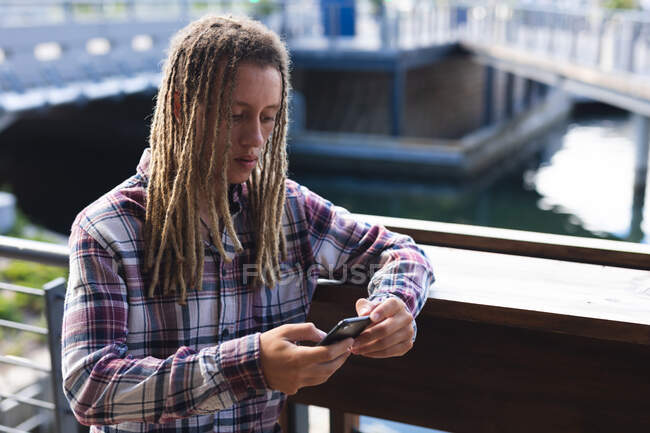 Homem de raça mista com dreadlocks em pé fora do café usando smartphone. nômade digital, para fora e sobre na cidade. — Fotografia de Stock