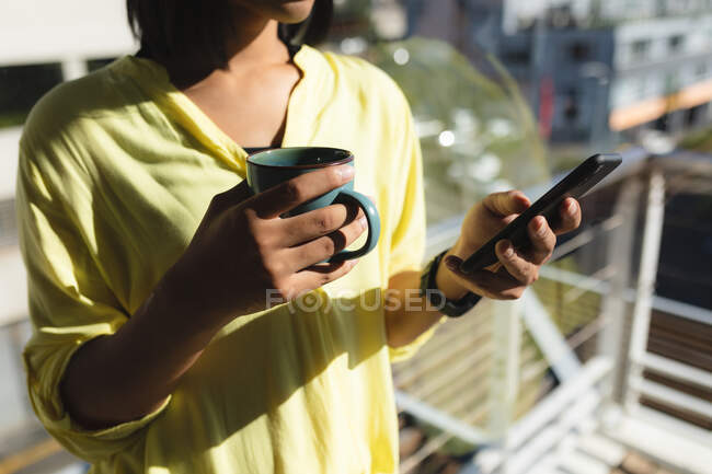 Midsection of mixed race transgender woman standing on roof terrace holding coffee using tablet. staying at home in isolation during quarantine lockdown. — Stock Photo