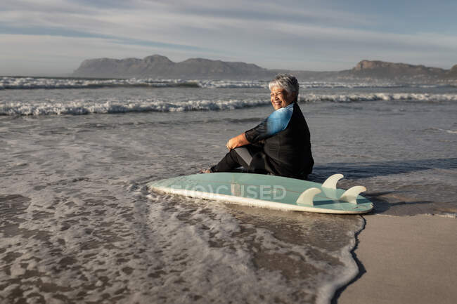 Eine ältere afrikanisch-amerikanische Frau mit Surfbrett lächelt, während sie am Strand sitzt. Reise Urlaub Ruhestand Lifestylekonzept — Stockfoto