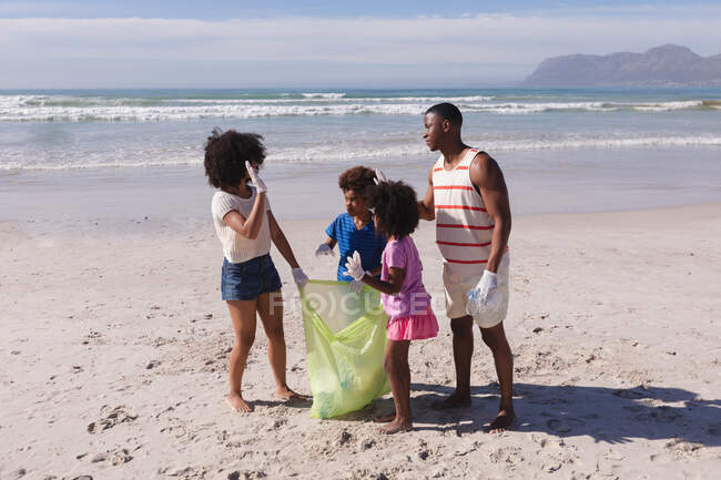 Afrikanisch-amerikanische Eltern mit zwei Kindern, die fünf Meter vom Strand entfernt Müll sammeln. Öko-Strandschutz für Familien. — Stockfoto