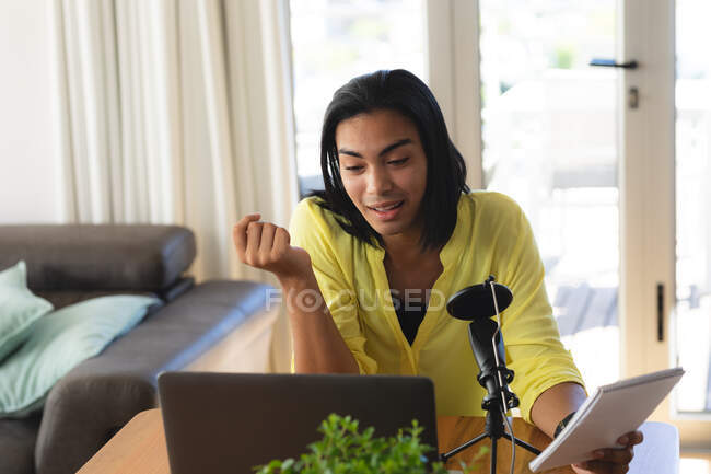 Mulher transgênero mista fazendo podcast usando microfone e laptop, conversando, segurando notas. ficar em casa em isolamento durante o confinamento de quarentena. — Fotografia de Stock