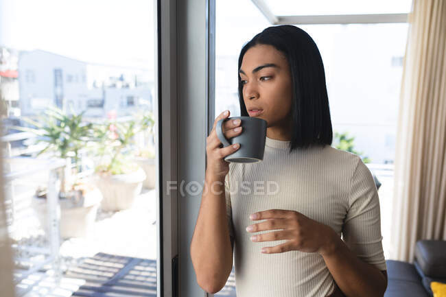 Mulher transgênero mista de pé olhando para fora da janela segurando xícara de café. ficar em casa em isolamento durante o confinamento de quarentena. — Fotografia de Stock