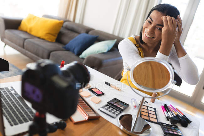 Happy mixed race transgender woman making vlog using laptop and camera putting on makeup. staying at home in isolation during quarantine lockdown. — Stock Photo