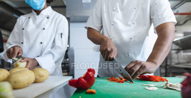 Sezione centrale di diversi chef professionisti gara preparare verdure indossando maschere viso. lavorando in una cucina ristorante occupato durante coronavirus covid 19 pandemia. — Foto stock
