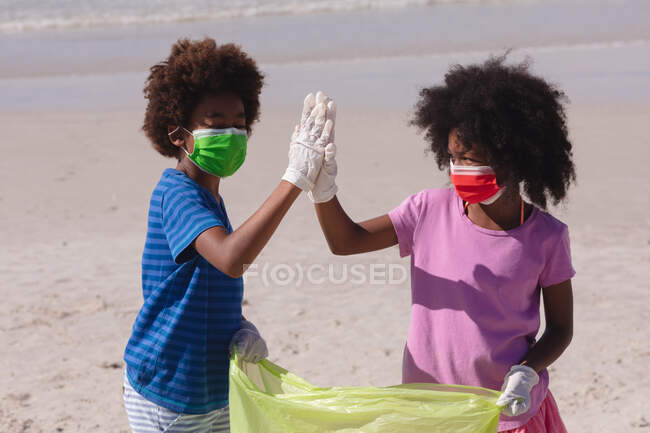 African american children wearing face masks collecting rubbish high fiving at the beach. family eco beach conservation during coronavirus covid 19 pandemic. - foto de stock