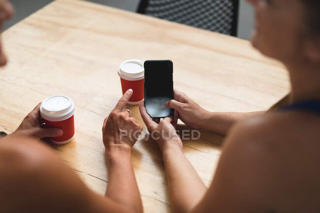 Mittelteil zweier glücklicher kaukasischer Frauen, die ihr Smartphone benutzen und Kaffee an einer Kletterwand trinken. Fitness und Freizeit im Fitnessstudio. — Stockfoto
