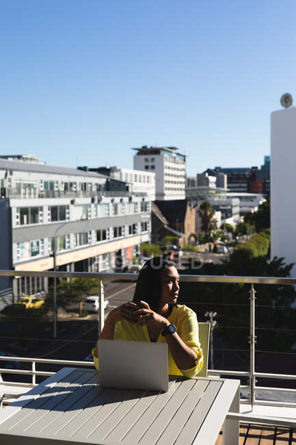 Mixed race transgender woman sitting at table on sunny roof terrace using laptop drinking coffee. staying at home in isolation during quarantine lockdown. — Stock Photo