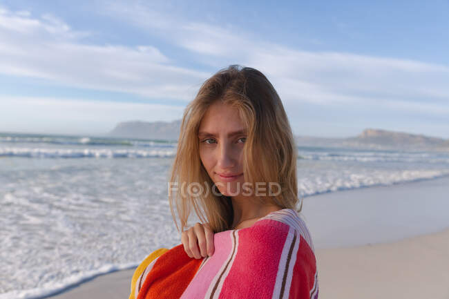 Donna caucasica avvolta da una coperta che guarda la telecamera e sorride alla spiaggia. Sano tempo libero all'aperto in riva al mare. — Foto stock