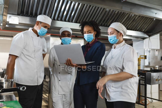 Diverso grupo de chefs profesionales que se reúnen con el gerente de cocina con máscaras faciales. trabajando en una ajetreada cocina de restaurante durante coronavirus covid 19 pandemia. - foto de stock