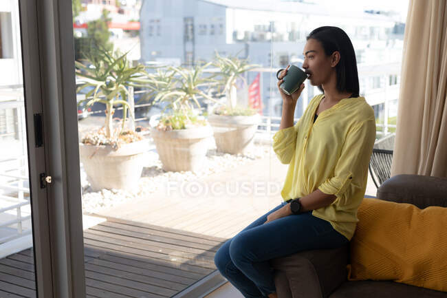 Sorrindo mestiço transgênero mulher sentada bebendo café, olhando para o terraço ensolarado. ficar em casa em isolamento durante o confinamento de quarentena. — Fotografia de Stock