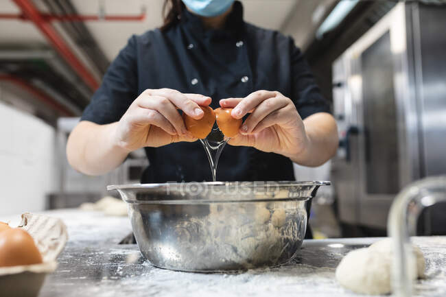 Sección media del chef profesional rompiendo huevos usando mascarilla. trabajando en una ajetreada cocina de restaurante durante coronavirus covid 19 pandemia. - foto de stock