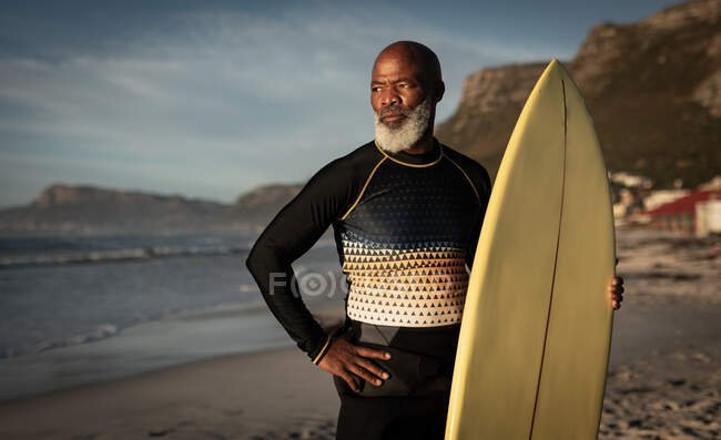 Портрет афроамериканського старшого чоловіка на пляжі, що тримає дошку для серфінгу з видом на море. здоров'я та благополуччя, активна пенсія . — стокове фото