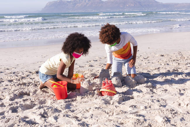 African american children wearing face masks playing with sand at the beach. family outdoor leisure time by the sea during coronavirus covid 19 pandemic. - foto de stock
