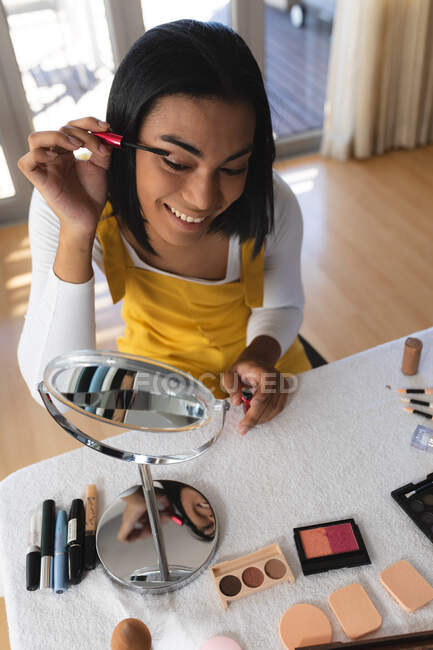 Mujer transexual de raza mixta sonriente sentada en la mesa mirando en el espejo poniéndose rímel. permanecer en casa aislado durante el bloqueo de cuarentena. - foto de stock