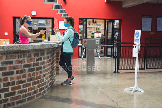 Femme réceptionniste et cliente portant des masques désinfectant les mains sur le comptoir au gymnase. fitness et temps libre à la salle de gym pendant coronavirus covid 19 pandémie. — Photo de stock