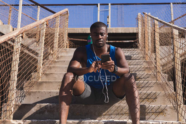 African american man exercising, wearing headphones, using smartphone on sunny day. healthy outdoor lifestyle fitness training. — Stock Photo