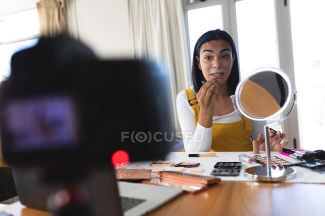 Смешанная расовая трансгендерная женщина делает видеоблог, используя ноутбук и камеру, накладывая макияж. оставаться дома в изоляции во время карантинной изоляции. — стоковое фото