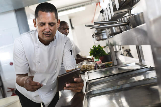 Mixed Race Profi-Koch schaut auf Bestellung und Tablet mit Kollegen im Hintergrund. Arbeit in einer belebten Restaurantküche. — Stockfoto