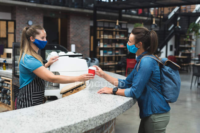 Dos mujeres caucásicas felices usando máscaras que pasan taza de café sobre el mostrador. fitness y tiempo libre en el gimnasio durante coronavirus covid 19 pandemia. - foto de stock