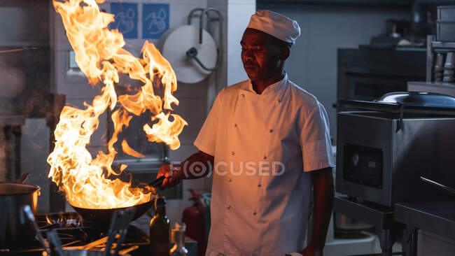 African american male professional chef flambeing dish in wok. working in a busy restaurant kitchen. — Stock Photo