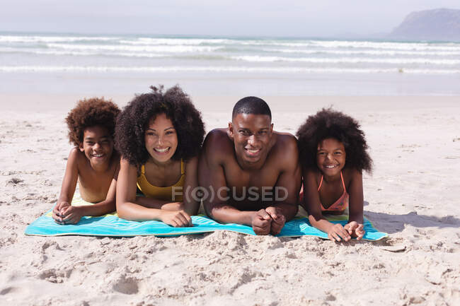 Retrato de pais afro-americanos e duas crianças deitadas em uma toalha na praia sorrindo. família tempo de lazer ao ar livre à beira-mar. — Fotografia de Stock