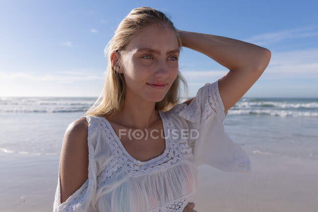 Улыбающаяся белая женщина, стоящая и трогающая волосы на пляже. здоровый отдых на открытом воздухе у моря. — стоковое фото