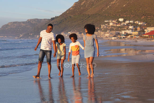 Smiling african american parents and two children walking and holding hands at the beach. healthy outdoor leisure time by the sea. — Photo de stock