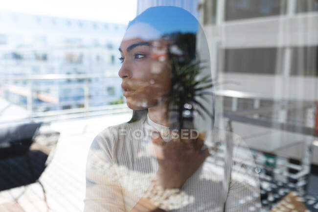 Змішана раса трансгендерна жінка, що стоїть у вікні, дивлячись на сонячну терасу на даху. перебування вдома в ізоляції під час карантину . — стокове фото