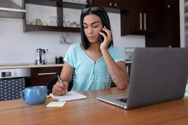 Mixed race transgender woman working at home using laptop talking on smartphone. staying at home in isolation during quarantine lockdown. — Stock Photo