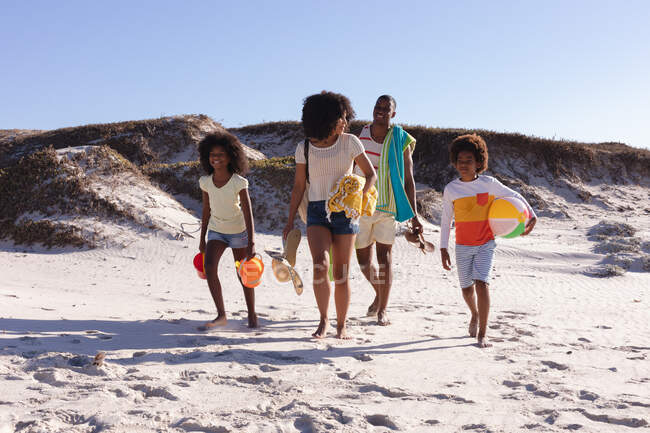 African american parents and two children holding beach accessories walking at the beach. family outdoor leisure time by the sea. — Fotografia de Stock