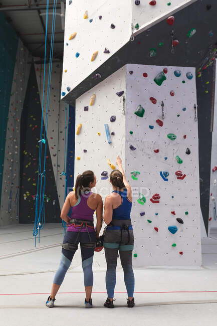 Zwei glückliche kaukasische Frauen unterhalten sich und bereiten sich auf einen Aufstieg an einer Indoor-Kletterwand vor. Fitness und Freizeit im Fitnessstudio. — Stockfoto