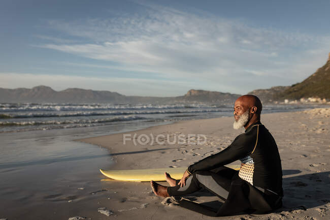 Африканський старший американець на пляжі сидить з дошкою для серфінгу, дивлячись на море. здоров'я і добробут, активний вихід на пенсію. — стокове фото