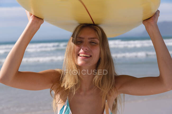 Усміхнена бікіні жінка носить жовту дошку для серфінгу на голові на пляжі. здоровий відпочинок на відкритому повітрі біля моря . — стокове фото