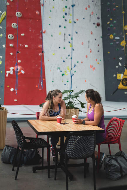 Duas mulheres caucasianas felizes conversando e bebendo café na parede de escalada interior. fitness e tempo de lazer no ginásio. — Fotografia de Stock