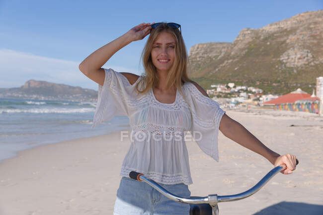 Mujer caucásica montando una bicicleta mirando a la cámara y sonriendo a la playa. Tiempo de ocio al aire libre saludable junto al mar. - foto de stock