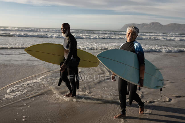 Happy senior african american couple carrying surf boards walking on the beach. travel vacation retirement lifestyle concept — Stock Photo