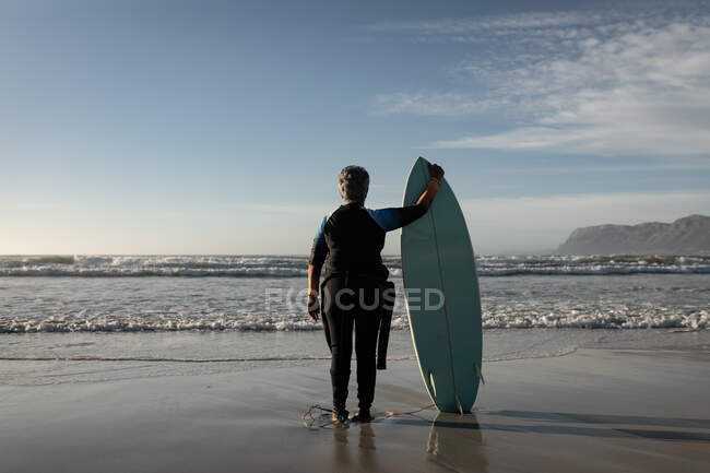Visão traseira da mulher americana africana sênior segurando prancha de surf em pé na praia. viagem férias aposentadoria estilo de vida conceito — Fotografia de Stock