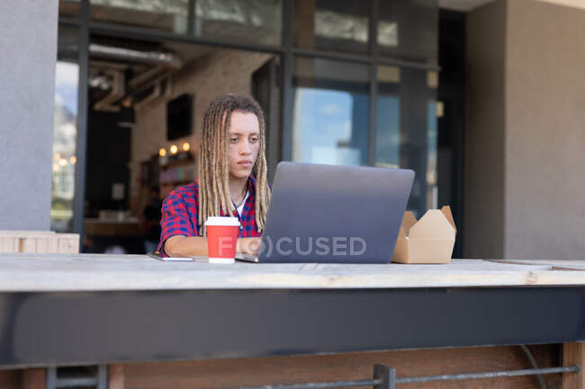 Homem de raça mista com dreadlocks sentado à mesa do lado de fora do café usando laptop. nômade digital, para fora e sobre na cidade. — Fotografia de Stock