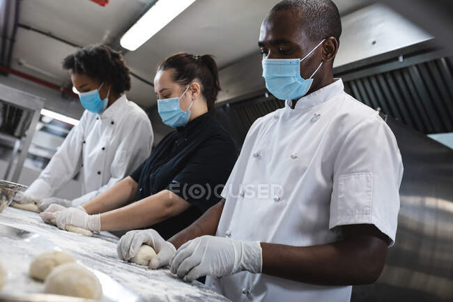 Chefs profesionales de raza mixta que preparan masa con guantes sanitarios y mascarilla facial. trabajando en una ajetreada cocina de restaurante durante coronavirus covid 19 pandemia. - foto de stock