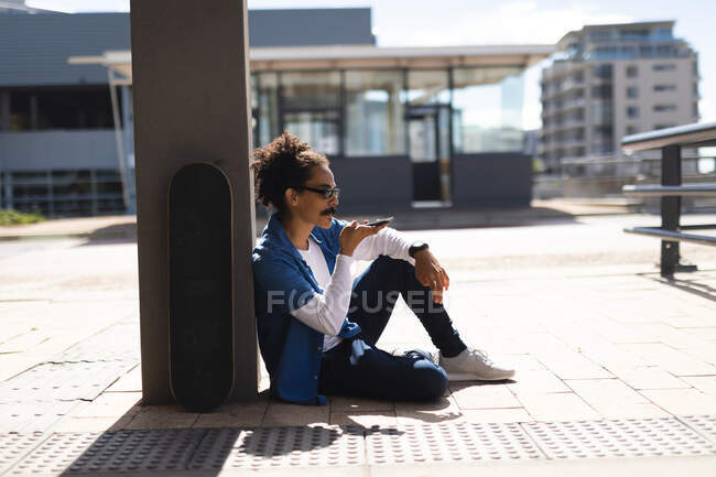 Misto raça macho com bigode sentado na rua com skate e falando no smartphone. nômade digital, para fora e sobre na cidade. — Fotografia de Stock