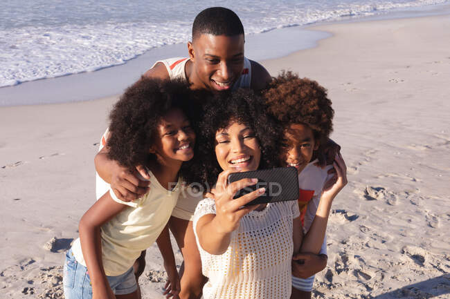 African american parents and two children taking a selfie with smartphone at the beach smiling. family outdoor leisure time by the sea. — Fotografia de Stock
