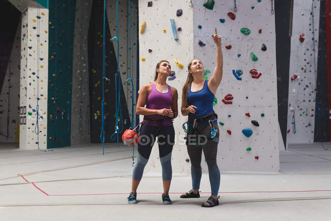 Dos mujeres caucásicas felices hablando y preparándose para una escalada en el muro de escalada interior. fitness y tiempo libre en el gimnasio. - foto de stock