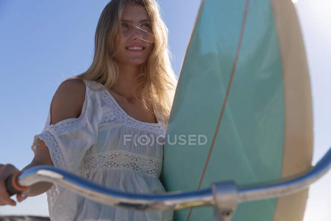 Mulher branca andando e carregando uma bicicleta e prancha de surf na praia. tempo de lazer ao ar livre saudável pelo mar. — Fotografia de Stock