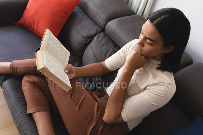 Смешанная расовая трансгендерная женщина отдыхает в гостиной, сидя на диване и читая книгу. оставаться дома в изоляции во время карантинной изоляции. — стоковое фото