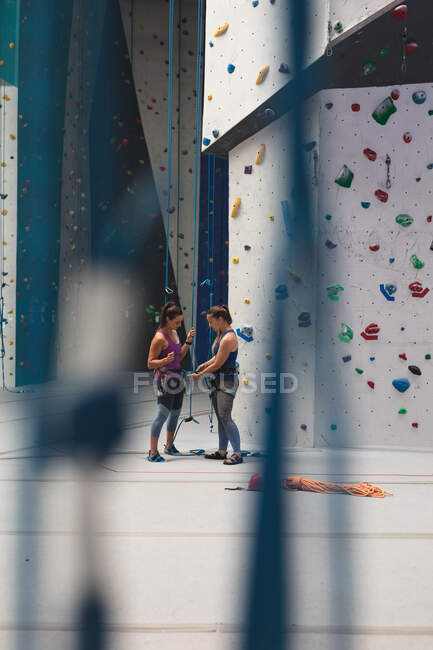 Kaukasische Instruktorin zeigt Frau, wie man an einer Indoor-Kletterwand ein Seil mit einem Gurtzeug verknotet. Fitness und Freizeit im Fitnessstudio. — Stockfoto