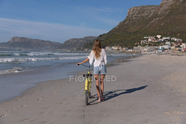 Задний вид кавказской женщины в белом топе и шорты, идущие на велосипеде по пляжу. здоровый отдых на открытом воздухе у моря. — стоковое фото