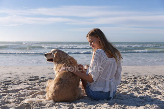 Кавказька жінка сидить на піску, чіпляючи собаку на пляжі. Здоровий вільний час на відкритому повітрі біля моря. — стокове фото