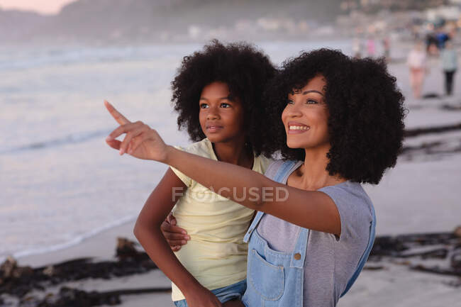 Smiling african american mother and daughter embracing at the beach, pointing. healthy outdoor leisure time by the sea. — Photo de stock