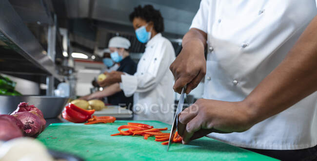 Різноманітні расові чоловічі та жіночі професійні кухарі готують овочі в масках для обличчя. робота на зайнятій кухні ресторану під час пандемії коронавірусу 19 . — стокове фото