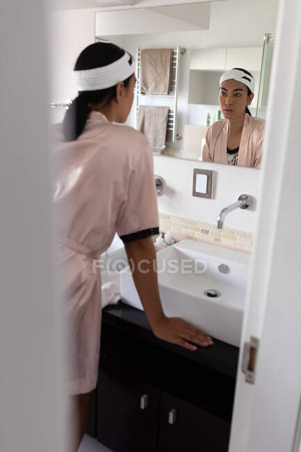 Transgender-Frau mit gemischter Rasse, die Stirnband und Robe trägt und in den Badezimmerspiegel blickt. Isolationshaft während der Quarantäne. — Stockfoto
